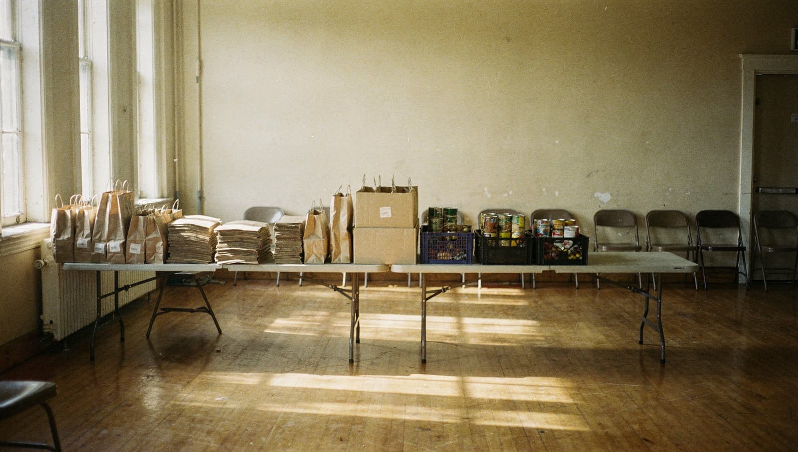 A community hall set up before a distribution: a long trestle table with packed paper bags, stacks of flat bags and crates of tins, empty chairs along the wall.