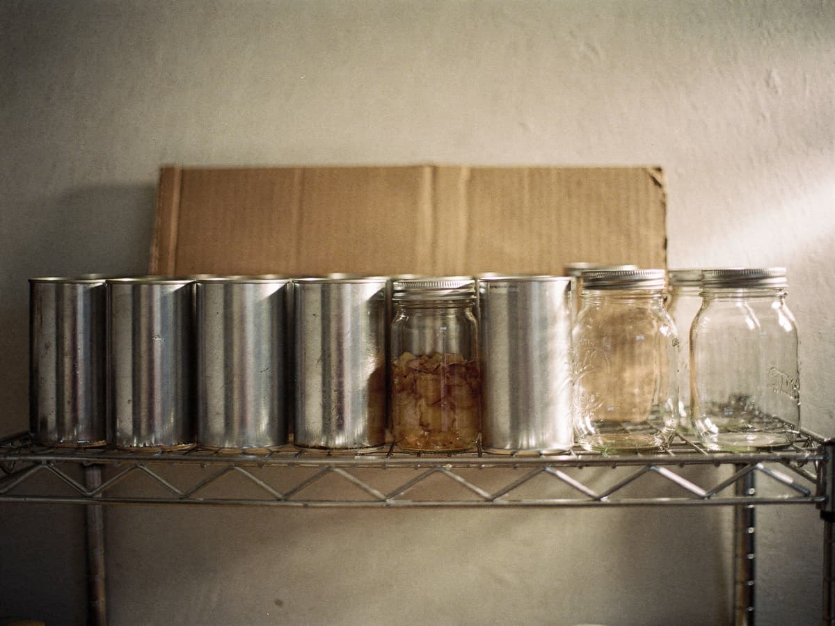 A row of plain tin cans and glass preserving jars on a wire shelf, lit from the side.